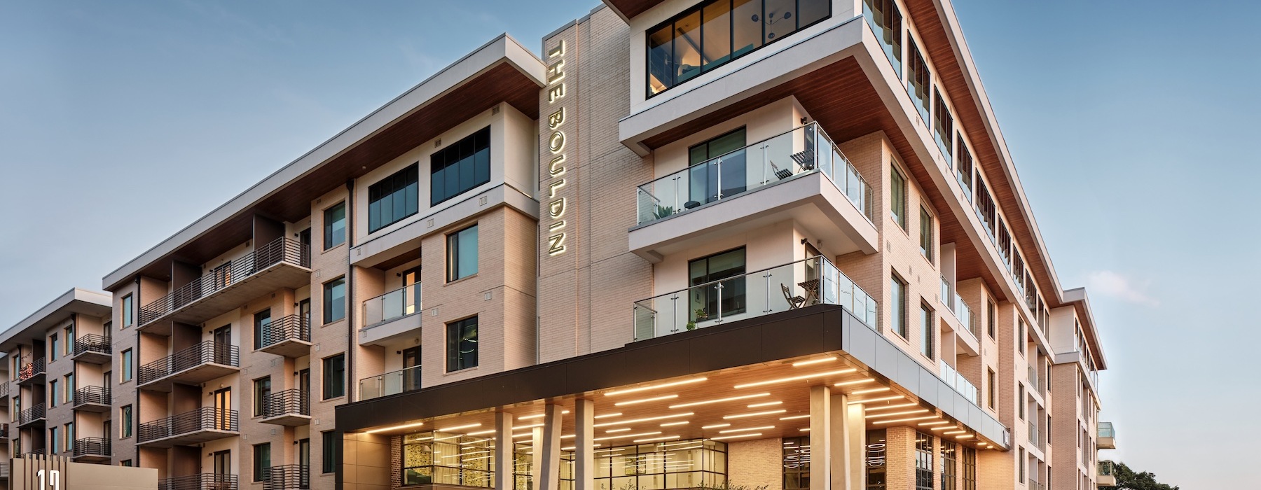 Exterior view of The Bouldin apartment community featuring contemporary architecture, private balconies, floor-to-ceiling windows, landscaped walkways, and a covered entrance with modern lighting.