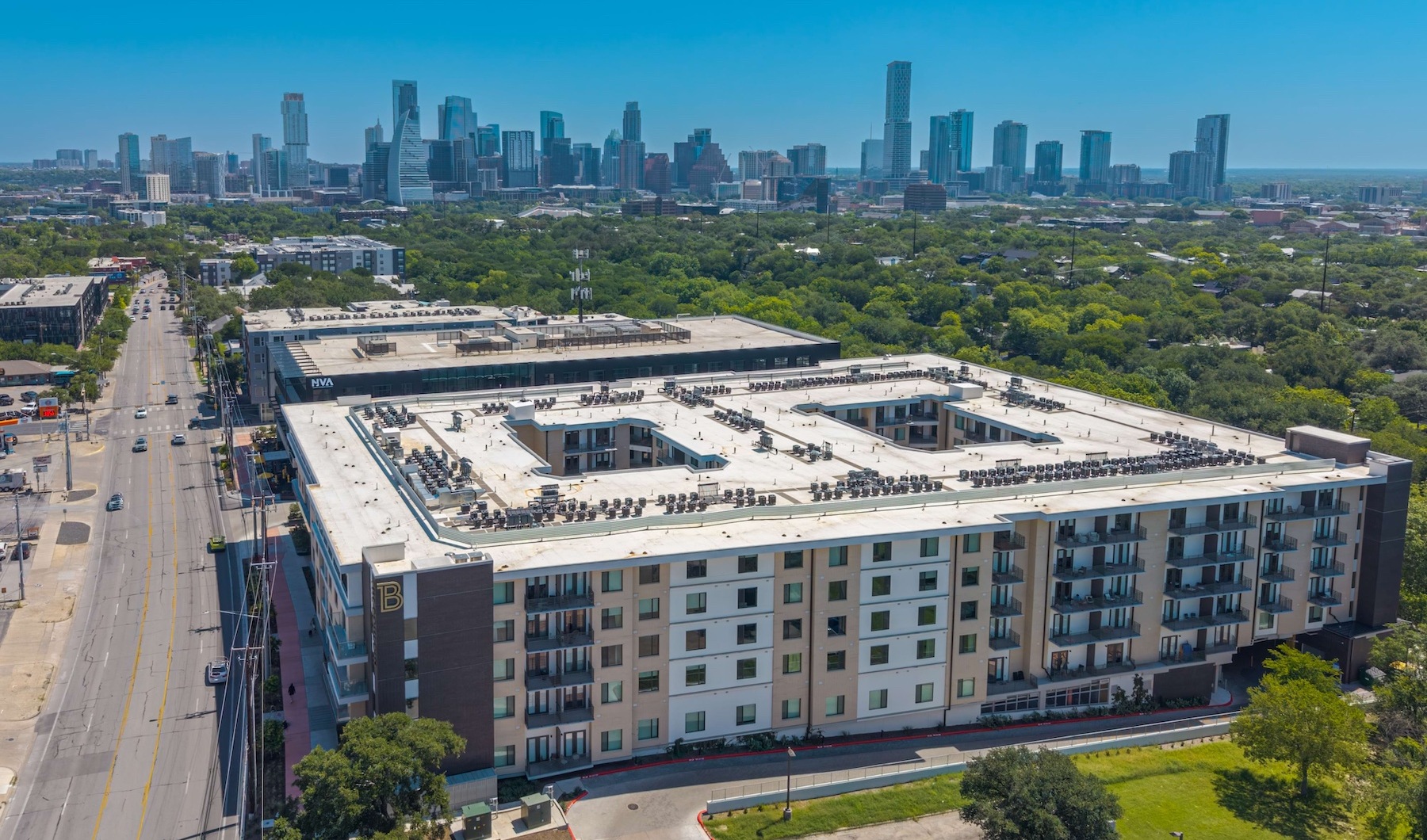 Aerial view of The Bouldin apartment community featuring a modern mid-rise residential building with private balconies, street-level retail, and panoramic views of downtown Austin and surrounding green space.