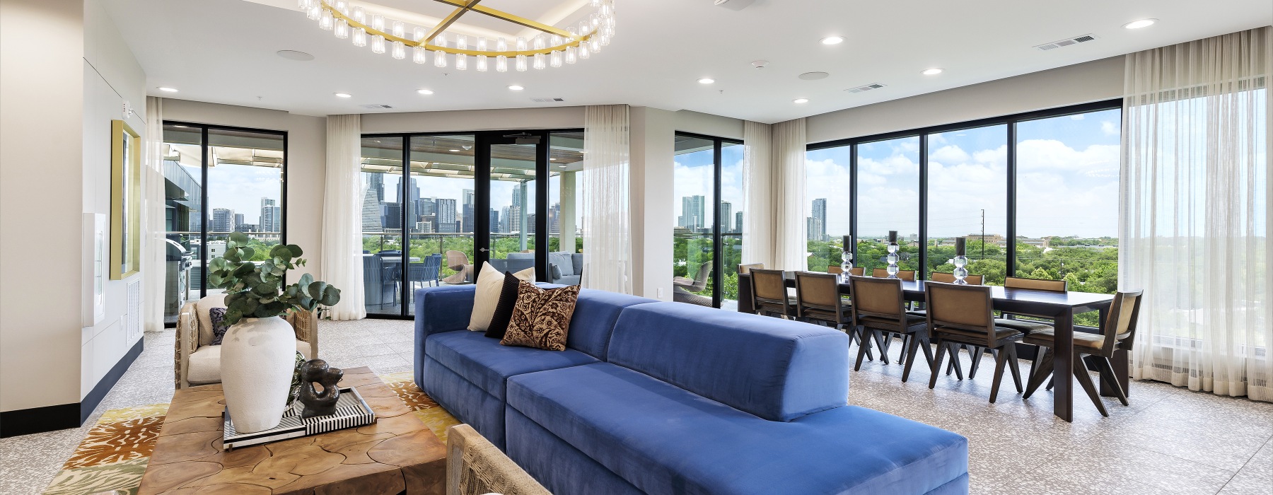 Resident lounge with a blue sectional sofa, seating area, dining table, large windows, and views of a city skyline and surrounding greenery.  For multifamily marketing, I would recommend using the Accessibility-Focused Alt Text on the website and reservin