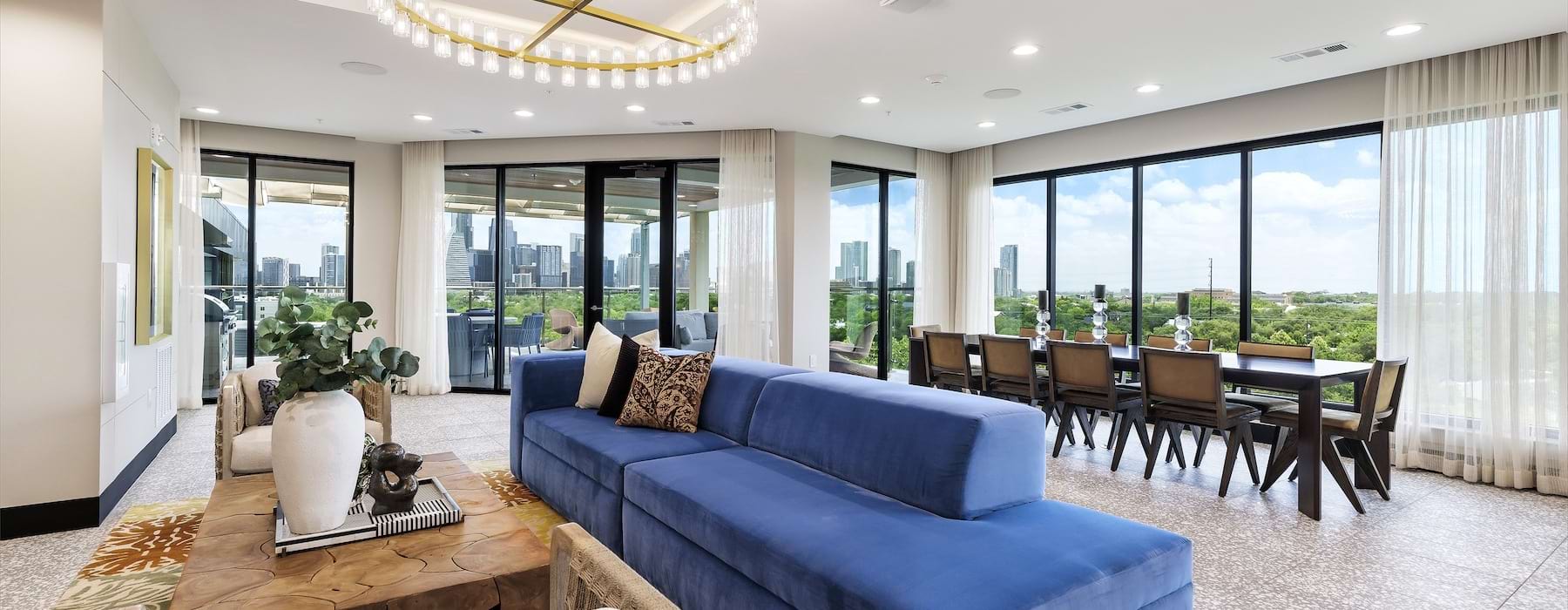 Resident lounge with a blue sectional sofa, seating area, dining table, large windows, and views of a city skyline and surrounding greenery.  For multifamily marketing, I would recommend using the Accessibility-Focused Alt Text on the website and reservin