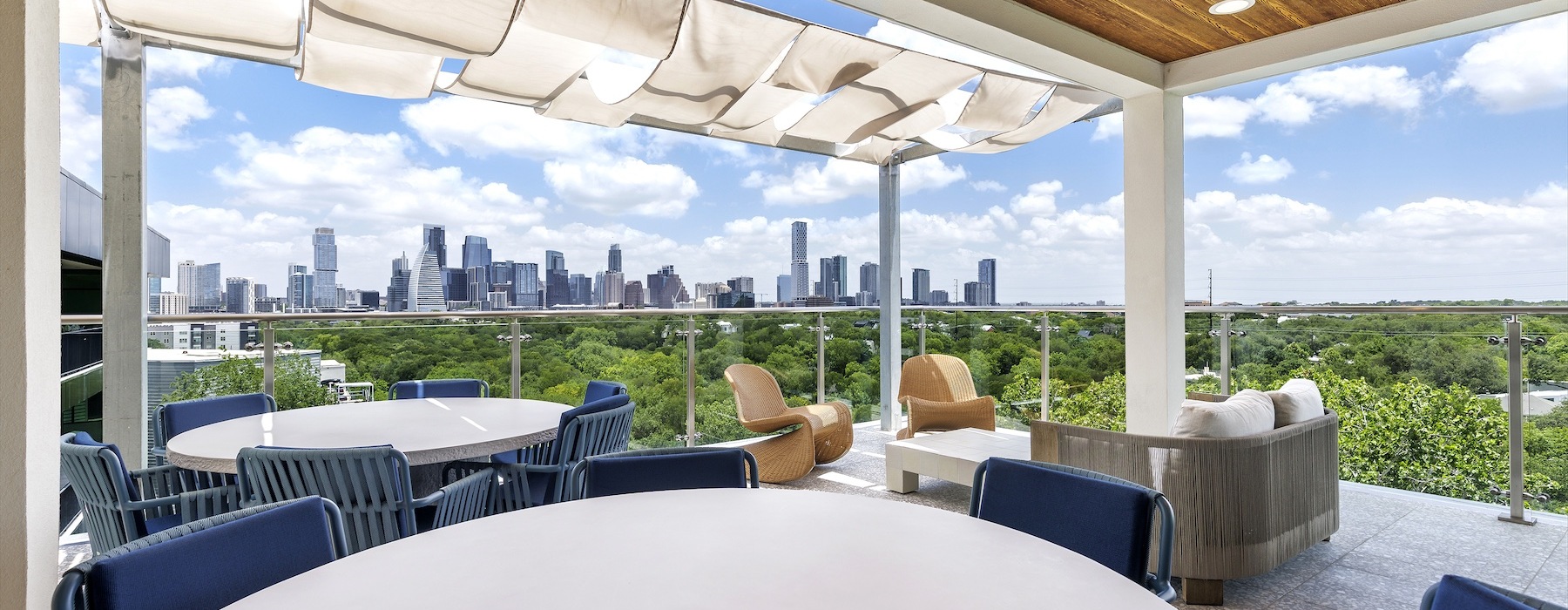 Rooftop terrace at The Bouldin featuring modern lounge seating, outdoor dining space, shaded canopy panels, and panoramic views of the Downtown Austin skyline above a lush greenbelt.